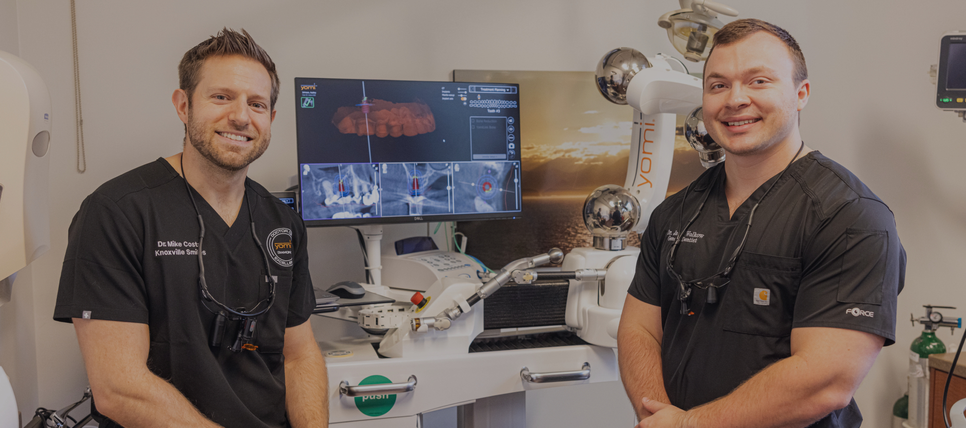 Two dental professionals smiling in modern dental operatory with equipment and monitors visible.