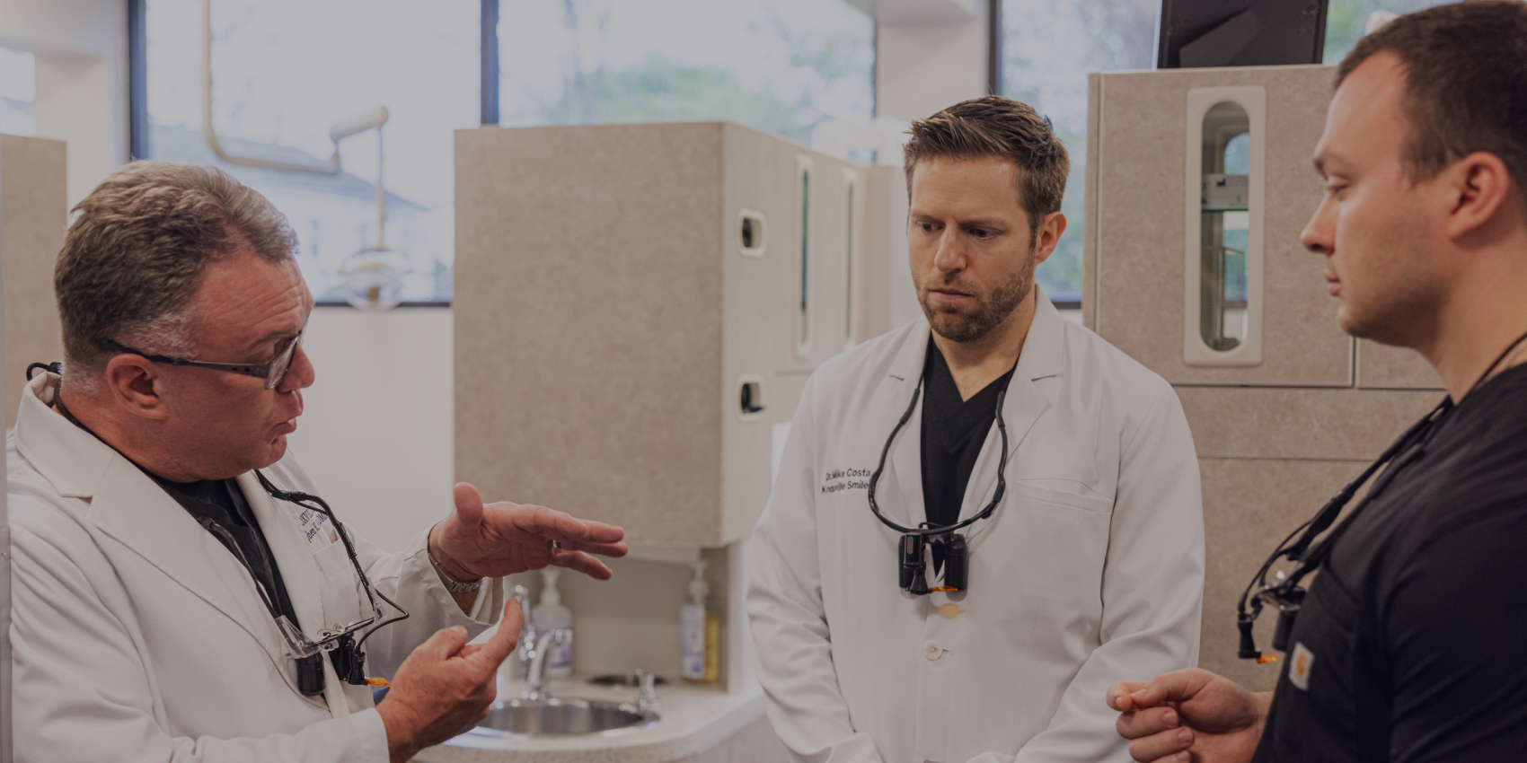 Three dental professionals discuss patient care in a modern dental office with treatment rooms visible in background.
