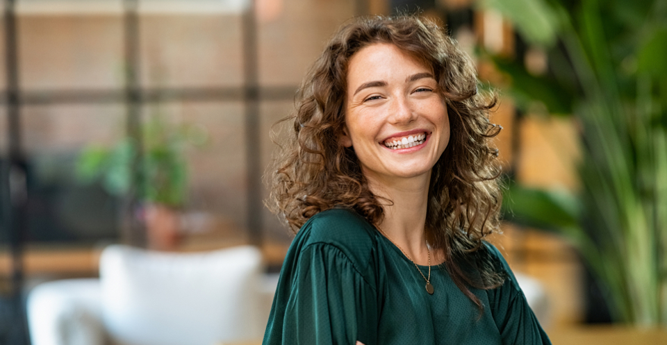 Woman with curly brown hair smiling at camera in modern dental office with plants