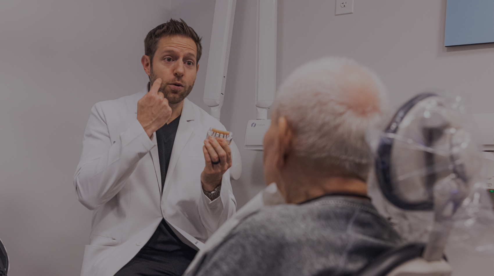 Male dentist in white coat gestures while consulting with elderly patient in dental office.