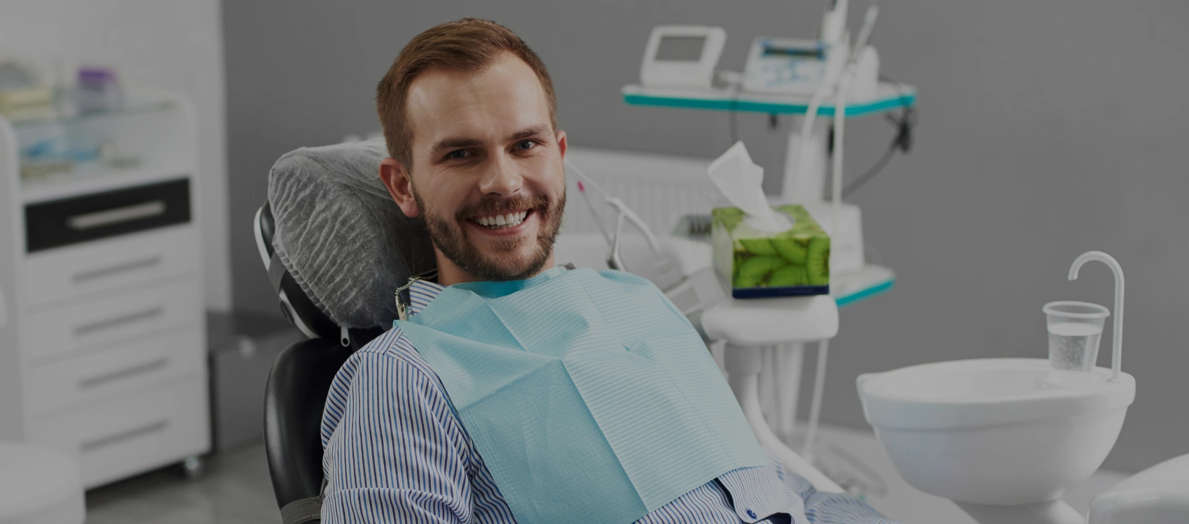 Male patient smiling in dental chair at modern dental office with equipment visible in background.