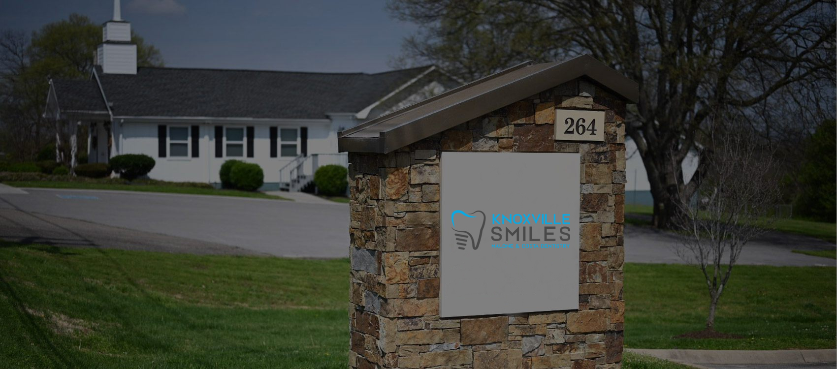 Dental office entrance sign with brick pillars and white building with steeple in background.