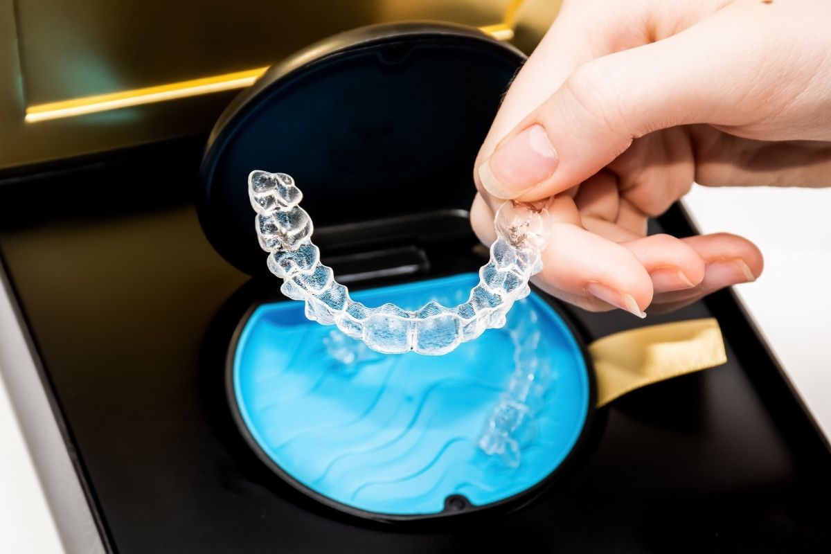 Person holding clear dental aligner over blue tray in dental office setting.